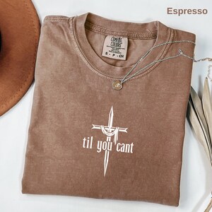 May include: Espresso-colored t-shirt with the text "til you cant" in white, featuring a cross design. The shirt has a crew neck and short sleeves. A silver necklace with a pendant is draped across the shirt.