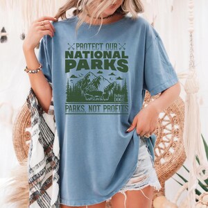 May include: A light blue t-shirt with a green graphic print that says "Protect Our National Parks, Defend Doge, Parks Not Profits". The graphic features a mountain range with trees and a bear.