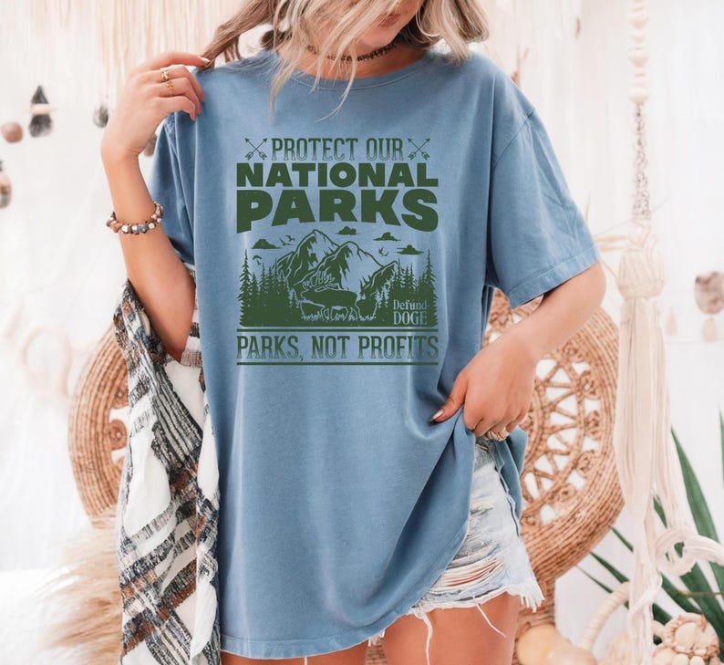 May include: A light blue t-shirt with a green graphic print that says "Protect Our National Parks, Defend Doge, Parks Not Profits". The graphic features a mountain range with trees and a bear.