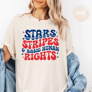 May include: Ivory-colored t-shirt with the words "STARS, STRIPES & BASIC HUMAN RIGHTS" in blue and red. The text is in a retro, bold font with star accents. The shirt is a casual, comfortable style.