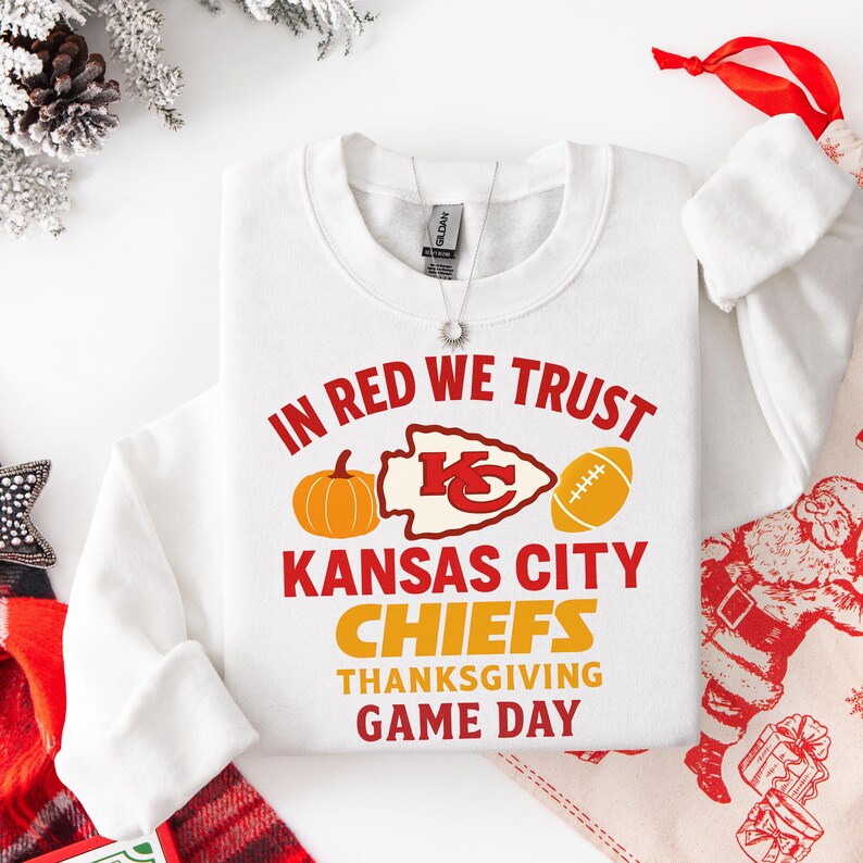 Puede incluir: Sudadera blanca con el texto "IN RED WE TRUST" y "KANSAS CITY CHIEFS THANKSGIVING GAME DAY" en rojo y naranja. El dise&ntilde;o incluye una calabaza, un bal&oacute;n de f&uacute;tbol americano y el logo de los Chiefs.