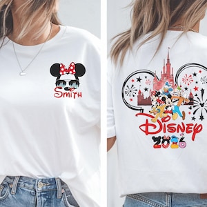 Pu&ograve; includere: T-shirt bianca con grafica di Minnie Mouse con occhiali da sole e il nome "Smith". Il retro della maglietta presenta un design Disney con personaggi, fuochi d'artificio e la scritta "Disney 2026".