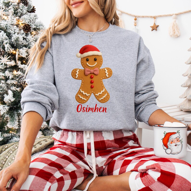 Op de afbeelding: Een grijze sweatshirt met een peperkoekmannetje met een kerstmuts en een rode vlinderdas. De naam "Osimhen" staat er in het rood onder. De persoon draagt een rood-witte geruite pyjamabroek en houdt een mok met een kerstmanontwerp vast.