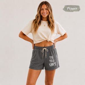 May include: A short-sleeved cream-colored t-shirt paired with charcoal gray shorts featuring an elastic waistband, a white drawstring, and the white text "TIL YOU CAN'T" printed on the lower right leg. The t-shirt is tucked at the waist.