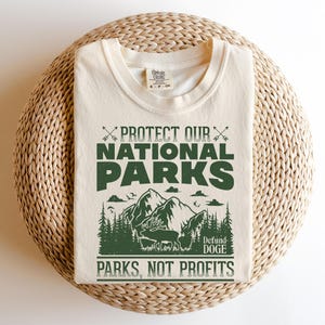 May include: A beige t-shirt with a green graphic print that says "Protect Our National Parks" with a mountain scene and the text "Defund Doge Parks, Not Profits".