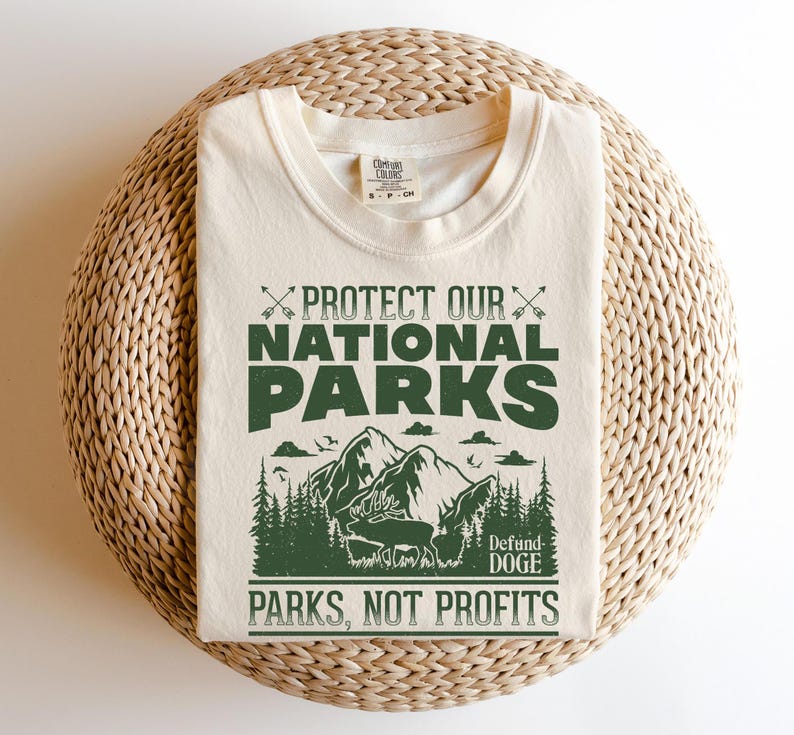 May include: A beige t-shirt with a green graphic print that says "Protect Our National Parks" with a mountain scene and the text "Defund Doge Parks, Not Profits".