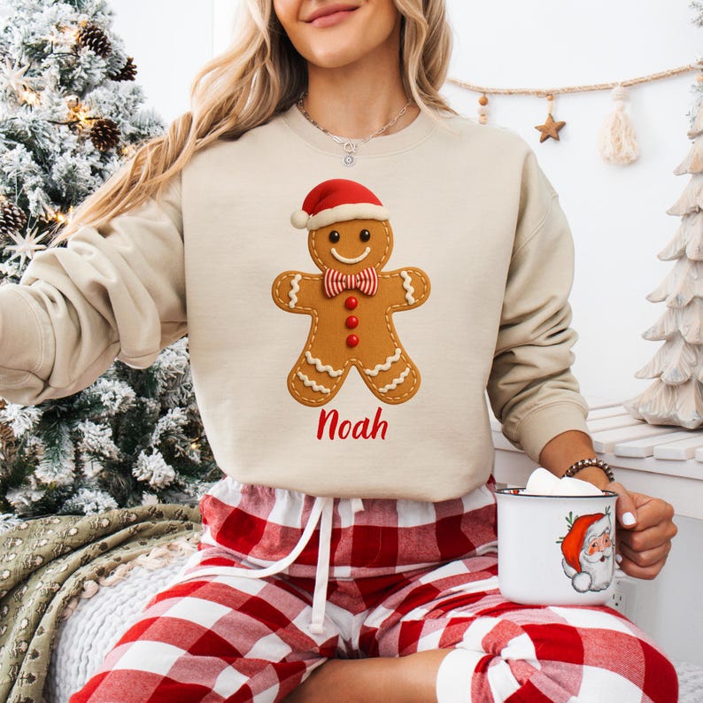 Op de afbeelding: Beige sweatshirt met een peperkoekmannetje met een kerstmuts en een strikje. De naam "Noah" staat er in het rood onder. De persoon draagt een rood-witte geruite pyjamabroek.