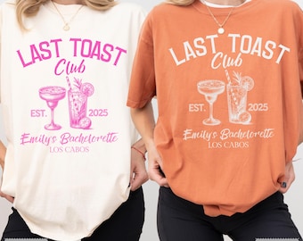 May include: Two t-shirts, one ivory and one yam-colored, feature the text "LAST TOAST Club" with a cocktail graphic, "EST. 2025," and "Emily's Bachelorette LOS CABOS." The shirts are casual and suitable for a bachelorette party.