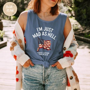 May include: A blue tank top with the text "I'M JUST MAD AS HELL" and an American flag graphic. The text "CAUS I LOVED THIS PLACE" is below the flag. The tank top is knotted at the waist.