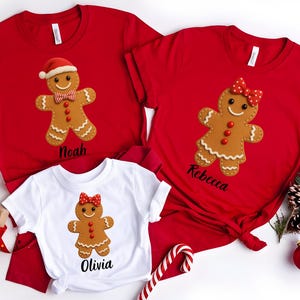 Op de afbeelding: Rode en witte kerst-T-shirts met peperkoekenmannetjesontwerpen. De rode shirts hebben een peperkoekenman met een kerstmuts en de naam "Noah", en een peperkoekenvrouw met een strik en de naam "Rebecca". Het witte shirt heeft een peperkoekenvrouw met de naam "Olivia".