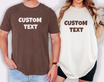 May include: Two t-shirts, one brown and one off-white, each with the words "CUSTOM TEXT" in bold, white letters. The brown shirt is paired with blue jeans, while the off-white shirt is paired with black shorts.