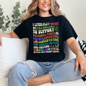 May include: A woman wearing a black t-shirt with the text "I literally want my tax dollars to support national parks, science, education, conservation, mental health and helping marginalized people. In case that wasn't clear."