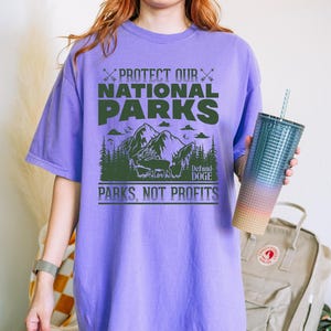 May include: Purple t-shirt with a green graphic that says "Protect Our National Parks" and "Defund Doge" with a mountain scene and the words "Parks, Not Profits".