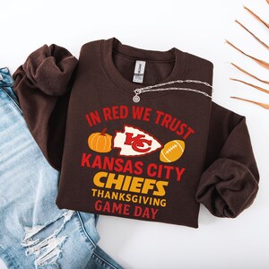 Puede incluir: Una sudadera marr&oacute;n con el texto "IN RED WE TRUST" y "KANSAS CITY CHIEFS THANKSGIVING GAME DAY" en rojo y amarillo. El dise&ntilde;o incluye una calabaza, un bal&oacute;n de f&uacute;tbol y el logotipo de los Chiefs. Una cadena plateada est&aacute; encima de la sudadera.