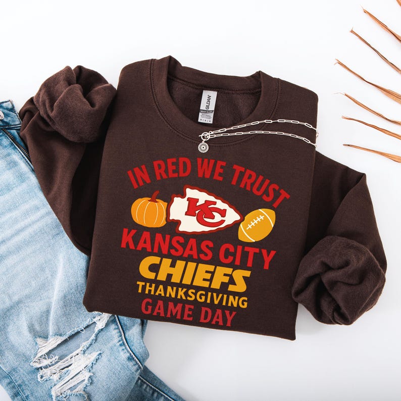 Puede incluir: Una sudadera marr&oacute;n con el texto "IN RED WE TRUST" y "KANSAS CITY CHIEFS THANKSGIVING GAME DAY" en rojo y amarillo. El dise&ntilde;o incluye una calabaza, un bal&oacute;n de f&uacute;tbol y el logotipo de los Chiefs. Una cadena plateada est&aacute; encima de la sudadera.
