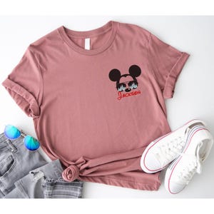 Pu&ograve; includere: Una maglietta rosa polveroso con una grafica di Topolino con occhiali da sole e il nome "Jackson" in corsivo rosso. La maglietta &egrave; abbinata a jeans blu, occhiali da sole e scarpe da ginnastica bianche con dettagli rossi e blu.