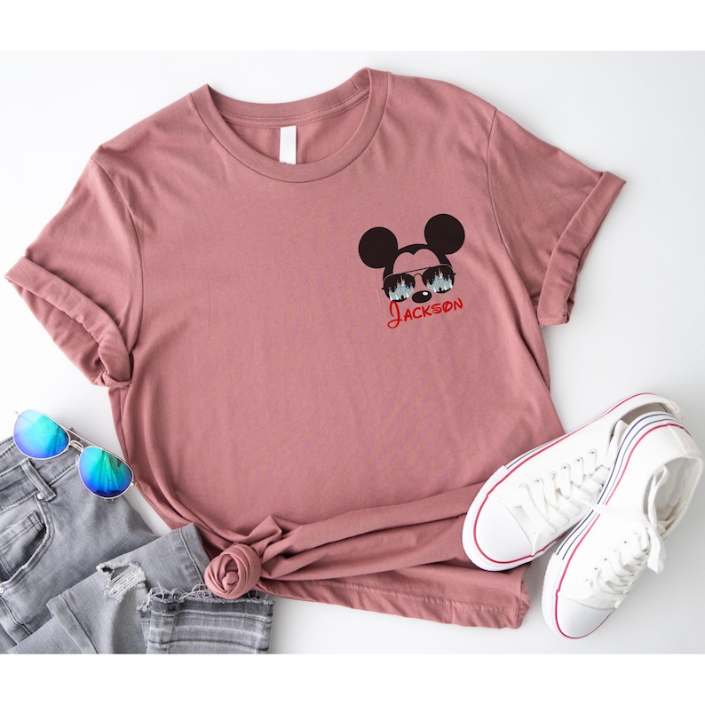 Pu&ograve; includere: Una maglietta rosa polveroso con una grafica di Topolino con occhiali da sole e il nome "Jackson" in corsivo rosso. La maglietta &egrave; abbinata a jeans blu, occhiali da sole e scarpe da ginnastica bianche con dettagli rossi e blu.