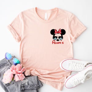 Pu&ograve; includere: T-shirt rosa chiaro con grafica di Minnie Mouse con occhiali da sole e fiocco rosso a pois bianchi. Il nome "Adam's" &egrave; scritto in rosso sotto. La maglietta &egrave; abbinata a jeans grigi, occhiali da sole e scarpe da ginnastica bianche.