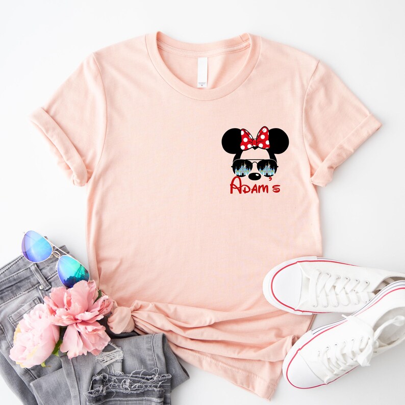 Pu&ograve; includere: T-shirt rosa chiaro con grafica di Minnie Mouse con occhiali da sole e fiocco rosso a pois bianchi. Il nome "Adam's" &egrave; scritto in rosso sotto. La maglietta &egrave; abbinata a jeans grigi, occhiali da sole e scarpe da ginnastica bianche.