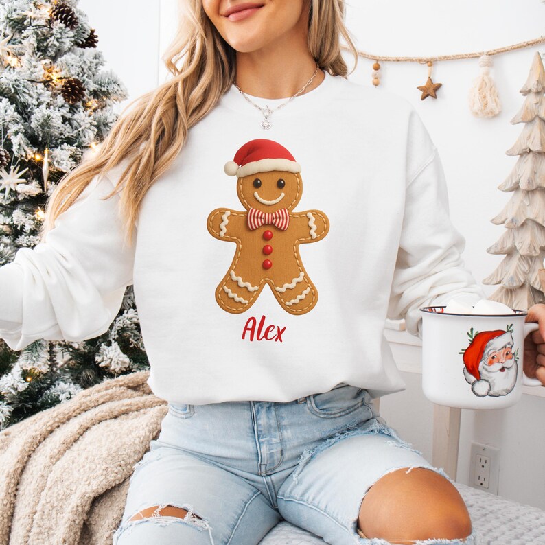 Op de afbeelding: Witte sweatshirt met een peperkoekmannetje met een kerstmuts en een strikje. Het peperkoekmannetje heeft een lachend gezicht en rode knopen. De naam "Alex" is in rode letters gedrukt. Een mok met een kerstmanontwerp is ook zichtbaar.