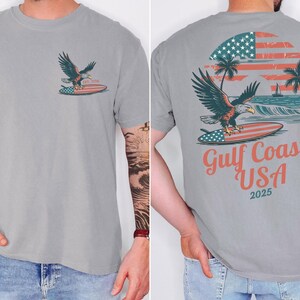 May include: A grey t-shirt featuring a graphic of an eagle on a surfboard with an American flag design. The back of the shirt displays the text "Gulf Coast USA 2025" with a sunset and palm trees.