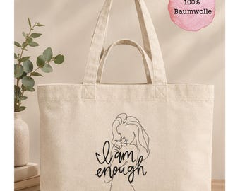 Könnte beinhalten: Eine beigefarbene Tragetasche aus Segeltuch mit zwei Henkelpaaren, bedruckt mit einer schwarzen Strichzeichnung einer Person und dem Text "I am enough". Ein rosa Kreis in der oberen rechten Ecke enthält den Text "100% Baumwolle".