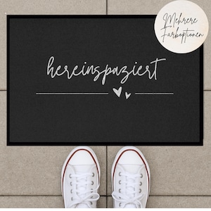 May include: Black welcome mat with white text that says "herein spaziert" with two small hearts underneath. The mat is on a gray surface with white sneakers in the foreground.