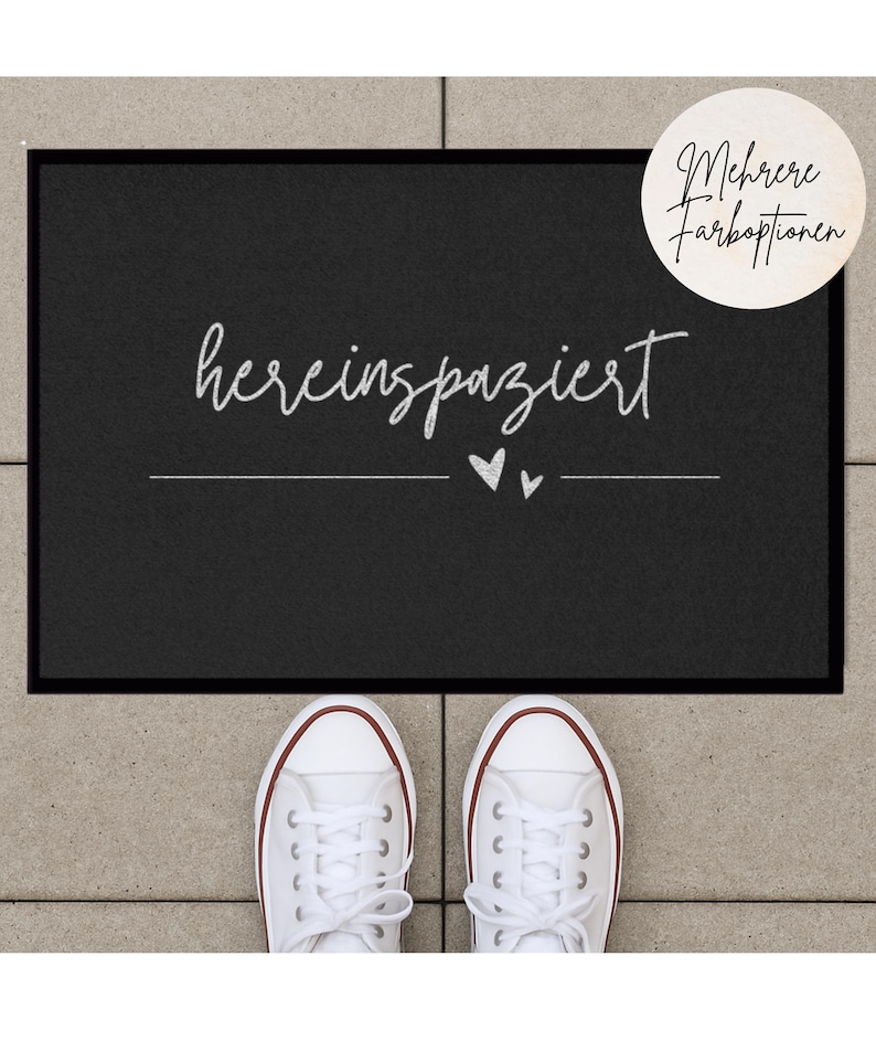 May include: Black welcome mat with white text that says "herein spaziert" with two small hearts underneath. The mat is on a gray surface with white sneakers in the foreground.