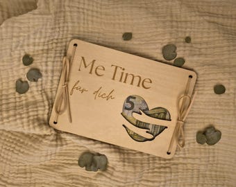 Könnte beinhalten: Eine rechteckige, helle Holzplatte mit der Gravur "Me Time" und "für dich". Ein herzförmiger Ausschnitt zeigt einen Fünf-Euro-Schein, wobei zwei Holzarme das Herz zu umarmen scheinen. Die Platte ist mit dünnen, beigen Kordeln zusammengebunden.