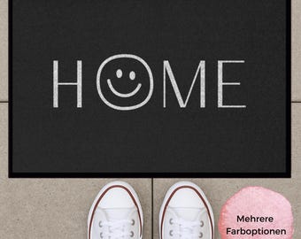 Könnte beinhalten: Eine rechteckige schwarze Fußmatte mit weißem Rand und dem Wort HOME in weißen Großbuchstaben, wobei der Buchstabe O durch ein weißes Smiley-Gesicht ersetzt ist. Die Matte liegt auf einem hellgrauen Fliesenboden.