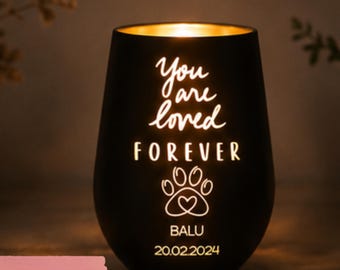 Könnte beinhalten: Ein mattschwarzer Kerzenhalter mit goldener Innenseite, der ein warmes Licht ausstrahlt. Auf der Vorderseite steht der Text "You are loved FOREVER" über einem Pfotenabdruck-Symbol mit Herz, gefolgt vom Namen "BALU" und dem Datum "20.02.2024" in Weiß.