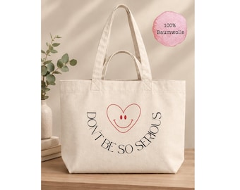 Könnte beinhalten: Eine cremefarbene Canvas-Tragetasche mit zwei Henkeln. Auf der Vorderseite befindet sich ein rotes, herzförmiges Smiley-Gesicht über dem schwarzen Text "DON'T BE SO SERIOUS" in einem Bogen. Ein rosa runder Aufkleber zeigt "100% Baumwolle".
