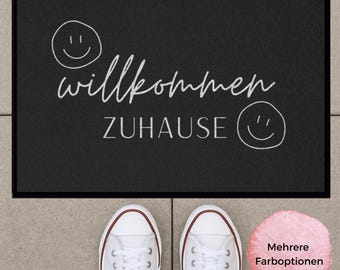 Könnte beinhalten: Eine rechteckige schwarze Fußmatte mit dem weißen Schriftzug "willkommen" in Schreibschrift und "ZUHAUSE" in Druckbuchstaben, ergänzt durch zwei handgezeichnete Smiley-Gesichter. Die Matte liegt oberhalb eines Paars weißer Sneaker.
