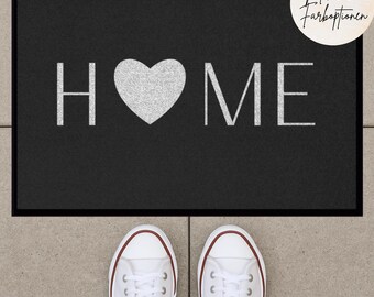 Può includere: Tappetino nero per porta con la scritta "HOME" in lettere bianche e un cuore argentato al centro. Il tappetino è su un pavimento piastrellato con un paio di scarpe da ginnastica bianche in primo piano.