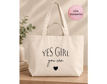 Könnte beinhalten: Eine cremefarbene Canvas-Einkaufstasche mit zwei Henkelpaaren und schwarzem Aufdruck mit dem Text YES GIRL you can sowie einem kleinen schwarzen Herzsymbol. Ein rosa kreisförmiges Abzeichen zeigt den Text 100% Baumwolle.