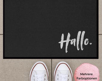Könnte beinhalten: Eine rechteckige schwarze Fußmatte mit dunklem Rand und dem Wort Hallo in weißer Pinselstrich-Schrift in der unteren rechten Ecke. Die Matte liegt über einem Paar weißer Sneaker auf einem hellgrauen, gefliesten Untergrund.