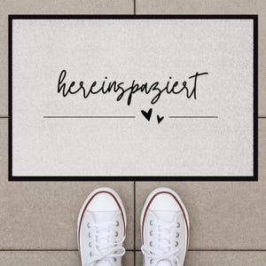 May include: A white doormat with black text that reads "hereinspaziert" with two small black hearts below the text.