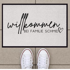 Peut inclure: Un paillasson blanc avec du texte noir qui dit "Willkommen bei Familie Schmid" avec un symbole de c&oelig;ur.