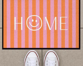Könnte beinhalten: Eine rechteckige Fußmatte mit vertikalen Streifen in abwechselnden Orange- und Rosatönen. Das Wort HOME ist in weißen Großbuchstaben in der Mitte aufgedruckt, wobei ein weißes Smiley-Gesicht den Buchstaben O ersetzt.