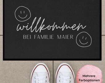 Könnte beinhalten: Eine rechteckige schwarze Fußmatte mit weißem Rand, die den Text "willkommen" in Schreibschrift, "BEI FAMILIE MAIER" in serifenloser Schrift sowie zwei handgezeichnete Smileys zeigt. Darunter befindet sich ein Paar weißer Sneaker.