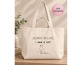 Könnte beinhalten: Eine beigefarbene Stofftasche mit dem schwarzen Aufdruck SORRY I'M LATE i saw a cat und einer einfachen Strichzeichnung einer sitzenden Katze. Die Tasche hat zwei Henkelpaare und ein rosa kreisförmiges Etikett mit 100% Baumwolle.