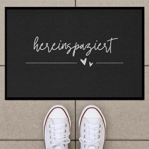 May include: Black welcome mat with the text "herein spaziert" in white lettering and two small white hearts.