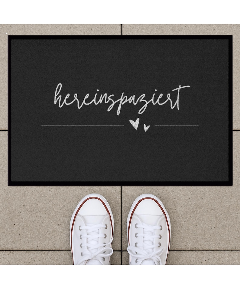 May include: Black welcome mat with the text "herein spaziert" in white lettering and two small white hearts.
