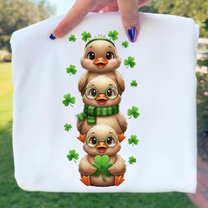 Puede incluir: Camiseta blanca con un diseño de tres patitos de dibujos animados apilados. El patito superior lleva una diadema verde con tréboles, el del medio tiene gafas y una bufanda verde, y el inferior sostiene un trébol. Tréboles verdes están dispersos.