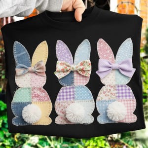 Puede incluir: Camiseta negra con tres diseños de conejos de patchwork. Cada conejo tiene orejas de patchwork coloridas, un lazo y una cola blanca y esponjosa. Los conejos están dispuestos en fila, creando un diseño festivo y caprichoso.