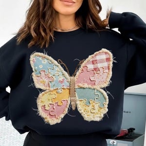 Puede incluir: Una sudadera azul marino con un gran diseño de mariposa. La mariposa está hecha de tela de patchwork en colores pastel, incluyendo rosa, amarillo, azul y cuadros rojos. Las alas de la mariposa tienen un diseño de piezas de rompecabezas.