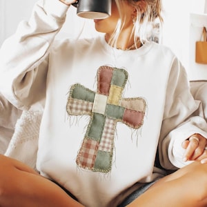 Puede incluir: Sudadera blanca con un diseño de cruz de retazos. La cruz está hecha de cuadrados de tela en tonos rosa, verde, amarillo y beige, con un estampado de cuadros. La cruz está cosida con un borde deshilachado.