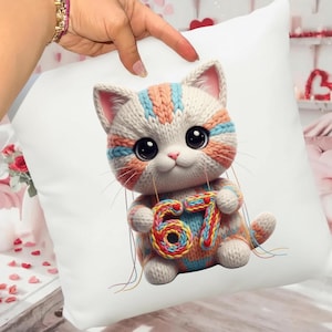 Puede incluir: Cojín cuadrado blanco con un diseño de gato tejido. El gato es en tonos blanco, melocotón y azul, sosteniendo el número colorido 67. El cojín tiene una apariencia suave y afelpada, ideal para la decoración del hogar.