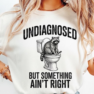 Puede incluir: Camiseta color crema con el texto negro "UNDIAGNOSED BUT SOMETHING AIN'T RIGHT". El diseño presenta un mapache en un inodoro en blanco y negro. Una camiseta gráfica humorística.
