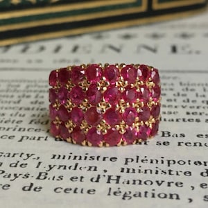 May include: A wide, gold-toned ring densely set with numerous round, vibrant red gemstones. The ring is displayed on a vintage paper background with faded text. The gemstones are closely packed, creating a continuous surface of color.